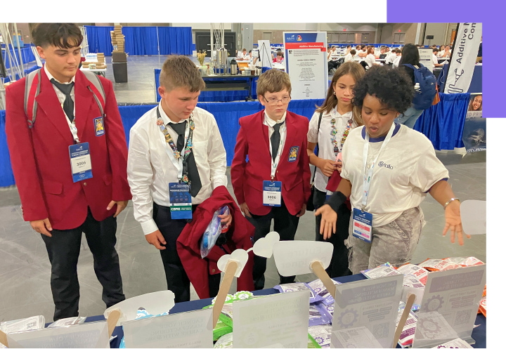 Five students in SkillsUSA competition attire gather around a table at a conference, with one student in a white polo demonstrating or explaining something to the group.