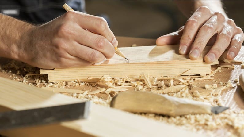 Close-up of a person's hands marking measurements on a wood plank at a workbench, with wood shavings scattered around.
