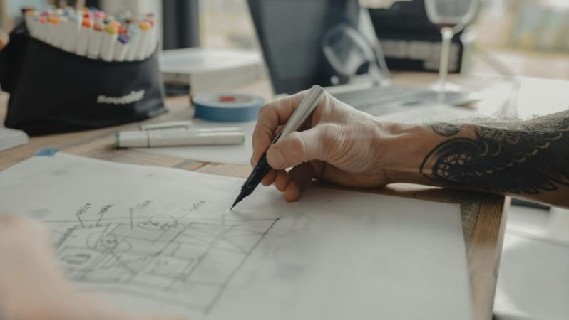 Close-up of a tattooed hand sketching a floor plan on paper with a pencil, next to markers and a laptop on a desk.