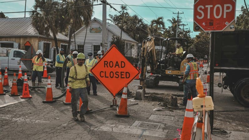 Road construction crew in safety vests working near a road closed sign, traffic cones, and an excavator.