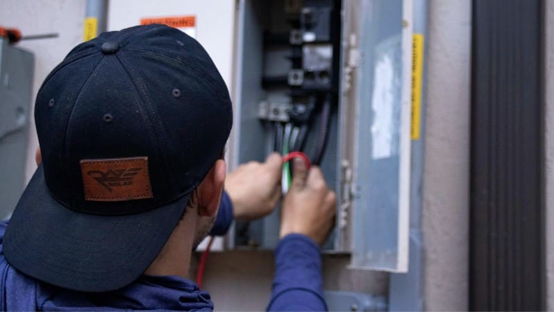 Electrician reaching into an open electrical panel to connect wires, wearing a baseball cap and long sleeve shirt.