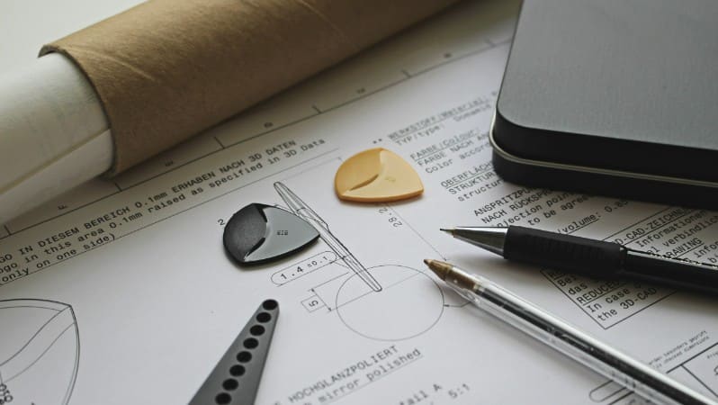 Technical drawing on a blueprint sheet surrounded by pens, a rolled paper tube, and small tool parts on a desk.