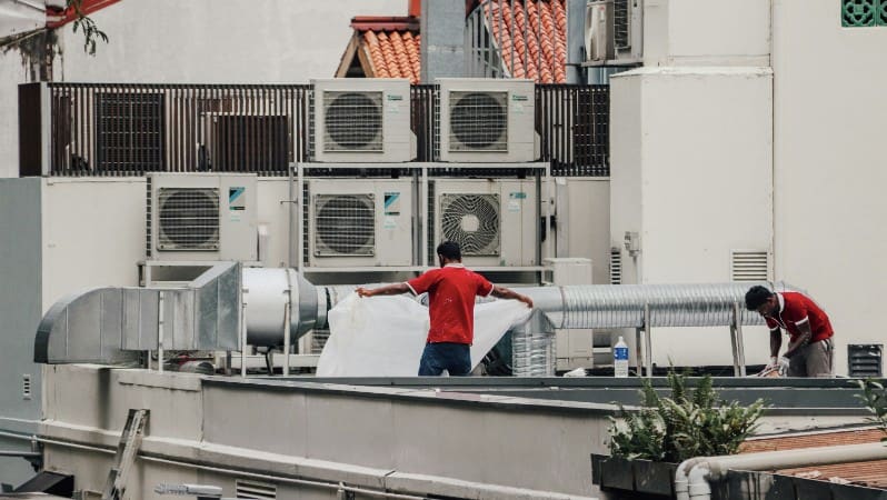 Two workers in red shirts servicing rooftop HVAC units and ductwork above a building.