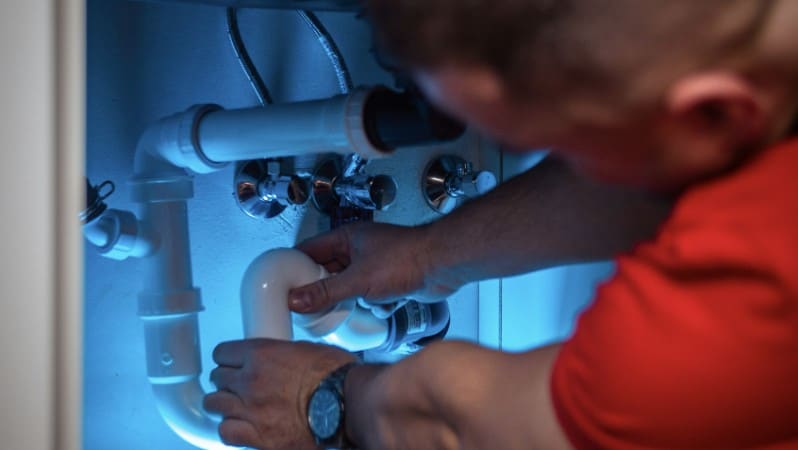 Close-up of a plumber's hands connecting PVC pipes under a sink, lit by a blue work light.