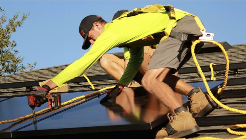 Worker in a safety harness kneeling on a rooftop, installing a solar panel with roofing tools and ropes nearby.