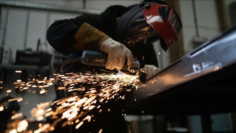 Construction worker in a face shield and gloves grinding a steel beam, sending off orange sparks in a metal shop.