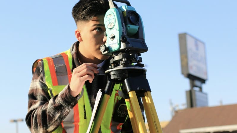 Worker in a hi-vis safety vest looking through surveying equipment on a tripod, with a signboard in the background.