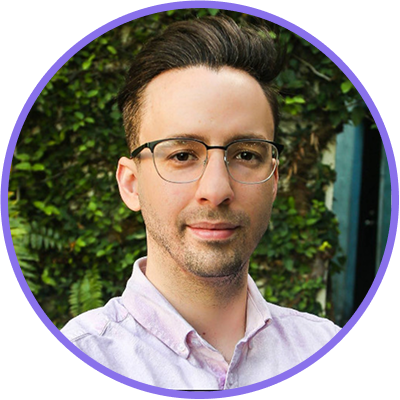 Headshot of Christian Gomez wearing glasses and a light pink button down shirt standing in front of green foliage.