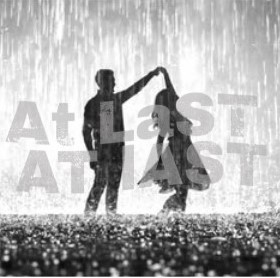 Silhouetted couple standing in heavy rain with one person lifting the other’s hand in a joyful pose as water splashes around their feet. Large faded text reading At Last overlays the scene, reinforcing a romantic and triumphant mood.