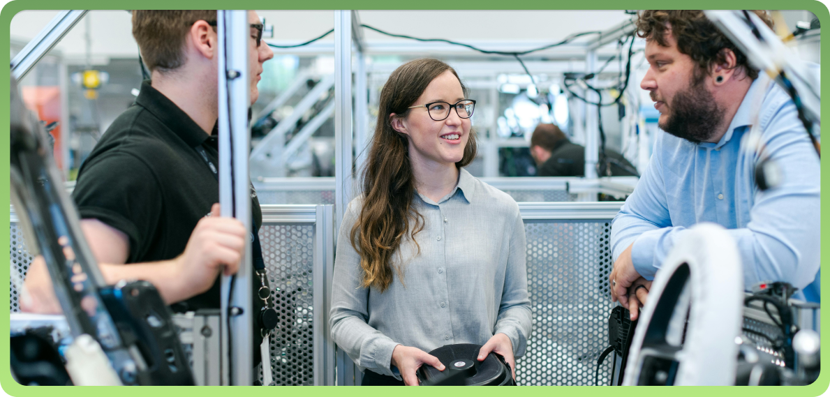 Three young professionals standing in a modern industrial workspace, smiling and talking among machinery. A woman in the center holds a component part, suggesting a collaborative engineering or internship setting.