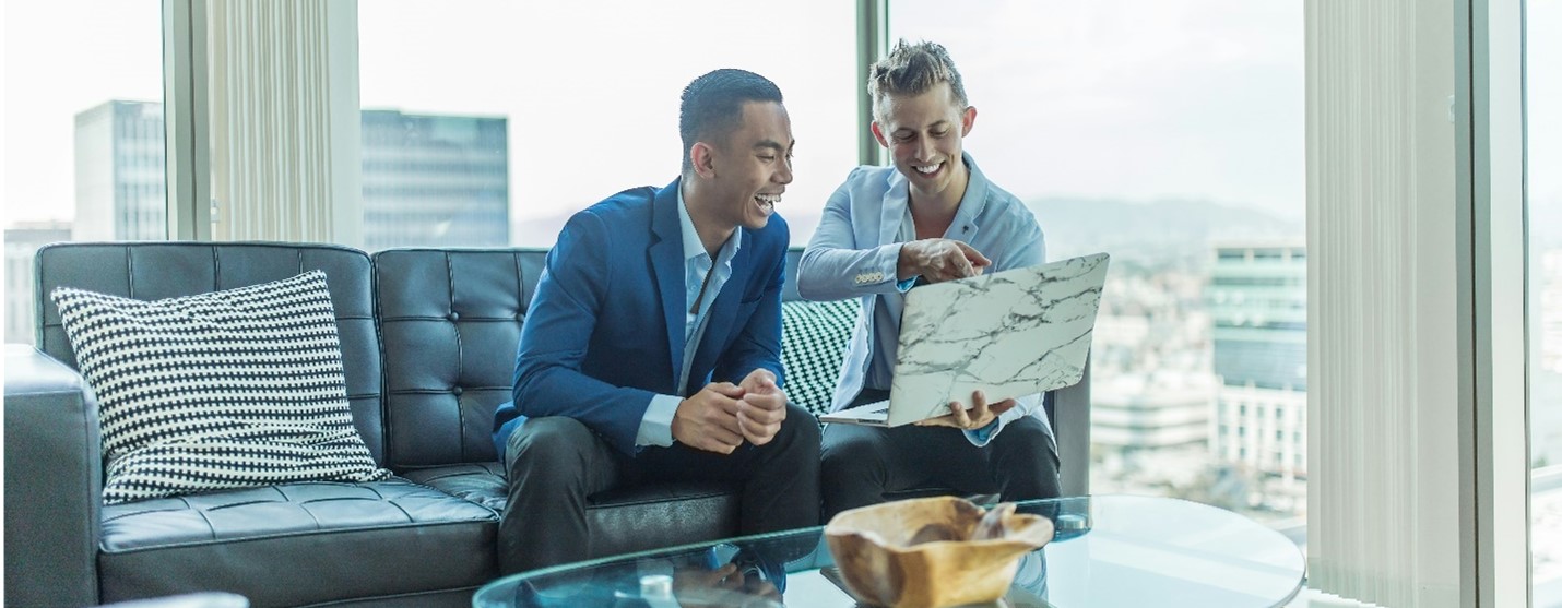 Two men sitting on a couch looking at one laptop and smiling, in an office setting