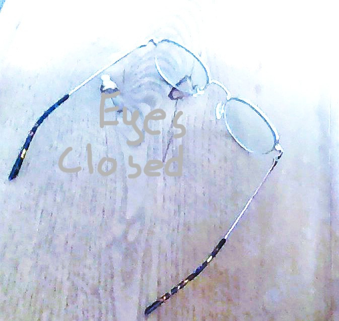 A pair of clear eyeglasses rests on a light wooden surface with soft cool toned lighting. Faint overlay text reads Eyes Closed, adding a reflective and introspective mood to the minimal composition.