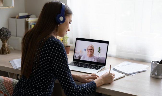 Back view of millennial female student in headphones note study distant with teacher or tutor on laptop. Young woman in earphones watch webinar, have online training or course with coach on computer.